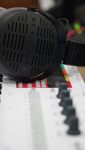 A pair of large, over-ear headphones rests on a colorful mixing console. The headphones are sleek and modern, with a visible brand logo and model number. The mixing console features numerous faders and knobs, with a color scheme that includes black, gray, and red accents.