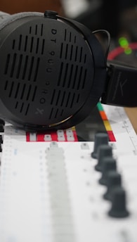A pair of large, over-ear headphones rests on a colorful mixing console. The headphones are sleek and modern, with a visible brand logo and model number. The mixing console features numerous faders and knobs, with a color scheme that includes black, gray, and red accents.