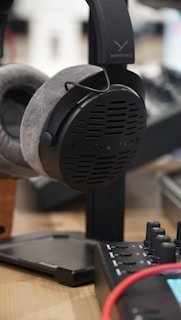 A close-up shot of a pair of Beyerdynamic headphones resting on a stand. The headphones have plush grey earcups and a black headband. In the foreground, part of a piece of audio equipment with buttons and dials, along with a red cable, can be seen.
