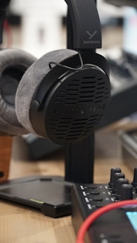 A close-up shot of a pair of Beyerdynamic headphones resting on a stand. The headphones have plush grey earcups and a black headband. In the foreground, part of a piece of audio equipment with buttons and dials, along with a red cable, can be seen.