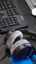 A pair of plush, over-ear headphones resting on a desk near a computer keyboard. The keyboard is black and has a wired connection. A blue notepad or document is partially visible beneath the headphones.