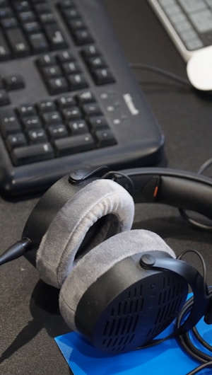 A pair of plush, over-ear headphones resting on a desk near a computer keyboard. The keyboard is black and has a wired connection. A blue notepad or document is partially visible beneath the headphones.
