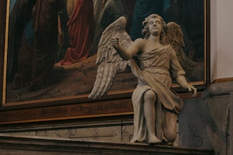 A detailed stone statue of an angel is positioned indoors, next to a large, colorful painting with religious imagery. The angel has large wings and is depicted in a kneeling pose, wearing a classical robe. The background painting features figures in robes, likely depicting a biblical scene.
