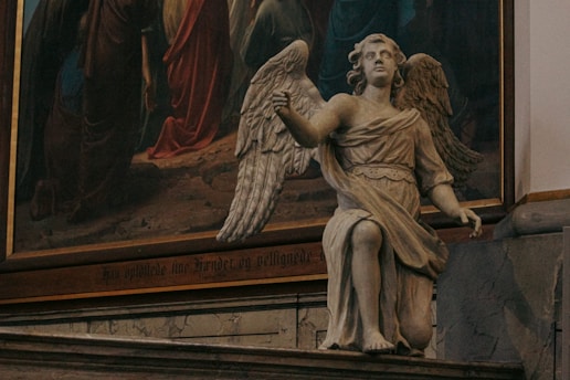 A detailed stone statue of an angel is positioned indoors, next to a large, colorful painting with religious imagery. The angel has large wings and is depicted in a kneeling pose, wearing a classical robe. The background painting features figures in robes, likely depicting a biblical scene.