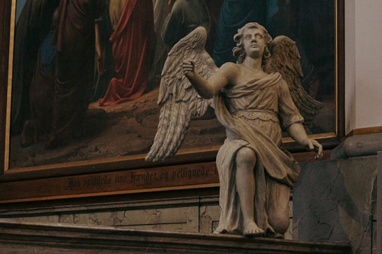 A detailed stone statue of an angel is positioned indoors, next to a large, colorful painting with religious imagery. The angel has large wings and is depicted in a kneeling pose, wearing a classical robe. The background painting features figures in robes, likely depicting a biblical scene.