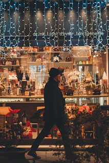 A person in a dark coat walks past a beautifully decorated shop window. The display is filled with assorted bottles, decorative wrapped gifts, and holiday-themed ornaments including small Christmas trees. Blue and white string lights hang from above, adding a festive atmosphere. The scene is captured during nighttime, evoking a warm and cozy holiday spirit.