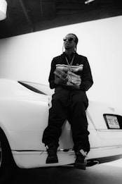 A person wearing sunglasses is sitting on the back of a white luxury car, holding a large stack of money. The person is dressed in dark clothing and is in a pose that suggests confidence. The setting appears to be indoors with a minimalistic background.