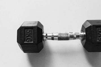 A single 5-kilogram hexagonal black dumbbell with a textured metal handle rests on a plain, light-colored background.
