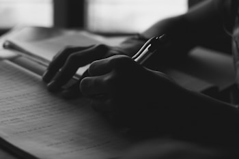 Silhouetted hands holding a pen, writing on lined paper with an open book visible in the background.