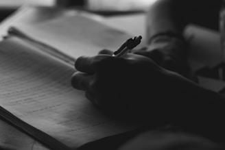 A silhouetted hand holding a pen is writing in a notebook. The focus is on the act of writing, with the background softly blurred.