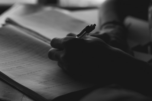 A silhouetted hand holding a pen is writing in a notebook. The focus is on the act of writing, with the background softly blurred.