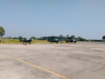 Several small aircraft are parked on a large open tarmac under a clear blue sky. There are a few trees and grass visible in the background. A person can be seen near the planes, possibly performing maintenance or inspection.
