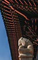 A detailed architectural roof design rich in colors and patterns with a focus on traditional elements. Below the roof, a stone statue resembling a lion is positioned prominently, displaying intricate carvings and a texture typical of historic sculptures.