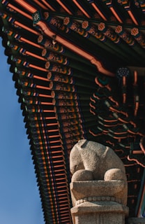 A detailed architectural roof design rich in colors and patterns with a focus on traditional elements. Below the roof, a stone statue resembling a lion is positioned prominently, displaying intricate carvings and a texture typical of historic sculptures.