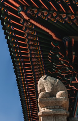 A detailed architectural roof design rich in colors and patterns with a focus on traditional elements. Below the roof, a stone statue resembling a lion is positioned prominently, displaying intricate carvings and a texture typical of historic sculptures.