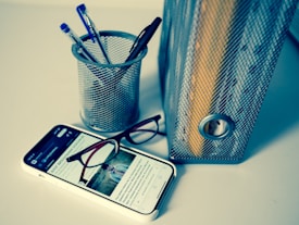 A desktop arrangement includes a metallic pen holder containing several pens, a smartphone displaying an article with glasses resting on it, and a metallic file holder with documents. The setting is minimalistic and organized.