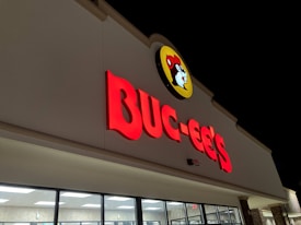 The image shows a brightly illuminated storefront sign for Buc-ee's featuring bold red lettering and a logo with a beaver face on a yellow background. The building exterior appears clean and modern with large windows below the sign, and the scene is set against a dark night sky.