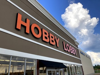 A large retail store with 'Hobby Lobby' prominently displayed in bold orange letters on the building facade. The design features a clean, modern look under a partly cloudy sky.