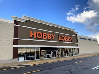 A large store building with a sign displaying 'HOBBY LOBBY' in bold orange letters. The store has a white facade with large windows at the front and a brown accent strip. The blue sky with white clouds is visible above the building.