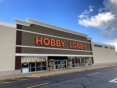 A large store building with a sign displaying 'HOBBY LOBBY' in bold orange letters. The store has a white facade with large windows at the front and a brown accent strip. The blue sky with white clouds is visible above the building.
