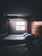 A dimly lit hospital room with two neatly made beds positioned next to a window. Light filters in from outside, casting faint shadows. There are curtains and medical equipment visible, including an IV pole, adding to the clinical atmosphere.