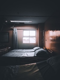A dimly lit hospital room with two neatly made beds positioned next to a window. Light filters in from outside, casting faint shadows. There are curtains and medical equipment visible, including an IV pole, adding to the clinical atmosphere.