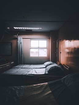 A dimly lit hospital room with two neatly made beds positioned next to a window. Light filters in from outside, casting faint shadows. There are curtains and medical equipment visible, including an IV pole, adding to the clinical atmosphere.