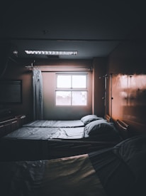 A dimly lit hospital room with two neatly made beds positioned next to a window. Light filters in from outside, casting faint shadows. There are curtains and medical equipment visible, including an IV pole, adding to the clinical atmosphere.