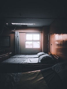 A dimly lit hospital room with two neatly made beds positioned next to a window. Light filters in from outside, casting faint shadows. There are curtains and medical equipment visible, including an IV pole, adding to the clinical atmosphere.