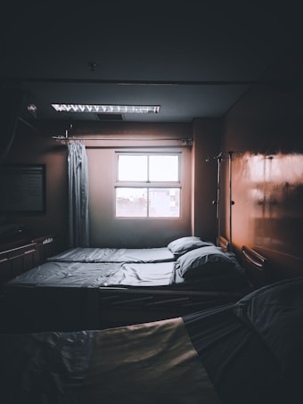 A dimly lit hospital room with two neatly made beds positioned next to a window. Light filters in from outside, casting faint shadows. There are curtains and medical equipment visible, including an IV pole, adding to the clinical atmosphere.