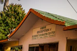 The image depicts a building with a sign in both Hindi and English that says 'Reception & Buddhist Information Counter'. It is part of the Mulagandha Kuty Vihara, Sarnath, Varanasi (UP). The roof is green with orange trim, and there are lush green trees in the background, along with a palm tree. The atmosphere appears sunny and peaceful.