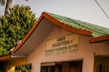 The image depicts a building with a sign in both Hindi and English that says 'Reception & Buddhist Information Counter'. It is part of the Mulagandha Kuty Vihara, Sarnath, Varanasi (UP). The roof is green with orange trim, and there are lush green trees in the background, along with a palm tree. The atmosphere appears sunny and peaceful.