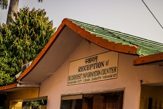 The image depicts a building with a sign in both Hindi and English that says 'Reception & Buddhist Information Counter'. It is part of the Mulagandha Kuty Vihara, Sarnath, Varanasi (UP). The roof is green with orange trim, and there are lush green trees in the background, along with a palm tree. The atmosphere appears sunny and peaceful.