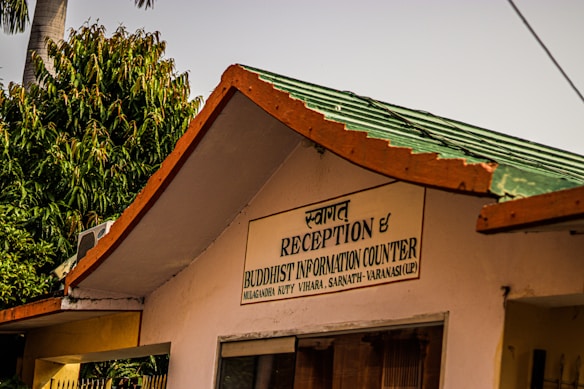 The image depicts a building with a sign in both Hindi and English that says 'Reception & Buddhist Information Counter'. It is part of the Mulagandha Kuty Vihara, Sarnath, Varanasi (UP). The roof is green with orange trim, and there are lush green trees in the background, along with a palm tree. The atmosphere appears sunny and peaceful.