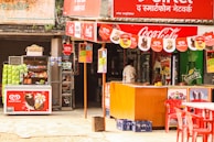 A small outdoor shop with colorful signage, featuring brands like Coca-Cola and Sprite. It has a display of various snacks and ice creams, with visible packets of chips and boxes of popsicles. There's a red counter and several red plastic chairs in front of the shop. A person is standing near the entrance, likely making a purchase.