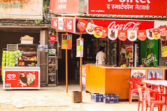A small outdoor shop with colorful signage, featuring brands like Coca-Cola and Sprite. It has a display of various snacks and ice creams, with visible packets of chips and boxes of popsicles. There's a red counter and several red plastic chairs in front of the shop. A person is standing near the entrance, likely making a purchase.