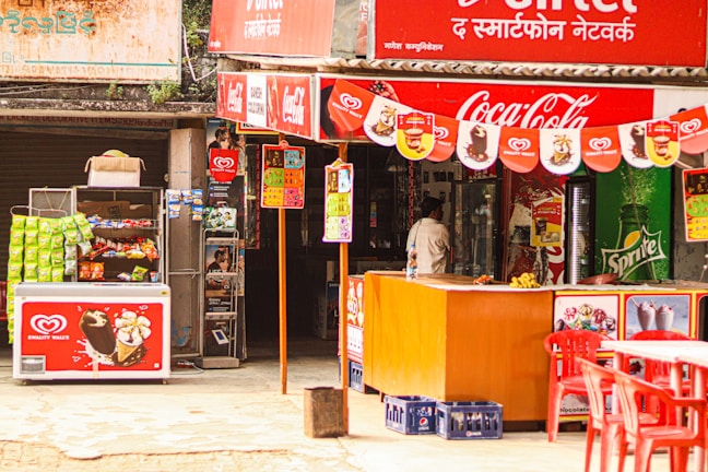 A small outdoor shop with colorful signage, featuring brands like Coca-Cola and Sprite. It has a display of various snacks and ice creams, with visible packets of chips and boxes of popsicles. There's a red counter and several red plastic chairs in front of the shop. A person is standing near the entrance, likely making a purchase.