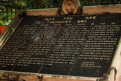A large stone plaque with engraved text in three columns, featuring Korean script. The plaque is mounted at an angle and appears to be situated outdoors with foliage and a wrought iron fence visible in the background. The plaque's surface is dark, with golden text that stands out clearly.