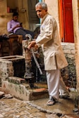 An older man in traditional attire operates a hand pump, with water dribbling into a stone basin in a rustic setting. Another man sits casually in the background, wearing a shirt and pants, focused on a phone. The scene conveys an everyday life moment in what appears to be an old house or small establishment.