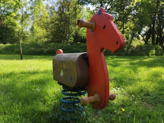 A red wooden rocking horse mounted on a metal spring is situated on a lush green grassy area under a canopy of trees. The background is filled with tall, green trees and dense foliage, creating a serene and natural atmosphere.