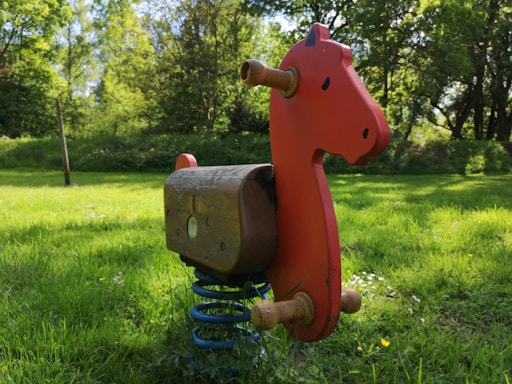 A red wooden rocking horse mounted on a metal spring is situated on a lush green grassy area under a canopy of trees. The background is filled with tall, green trees and dense foliage, creating a serene and natural atmosphere.