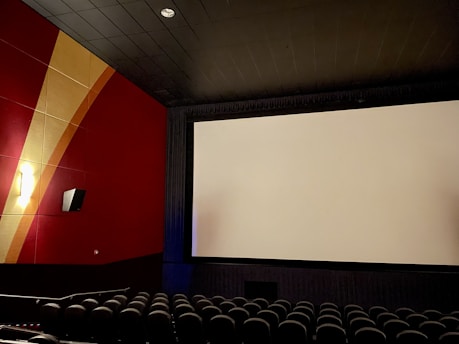 An empty movie theater featuring rows of black seats facing a large, blank cinema screen. The walls are decorated in a combination of red and gold tones with an abstract pattern. The ceiling is dark, with lighting providing a soft, ambient glow.