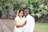 A couple is standing together in a garden, dressed in traditional attire. The woman is wearing a white saree with gold accents and has adorned her hair with flowers. The man is wearing a white shirt and is smiling warmly. They are embracing each other, expressing happiness.