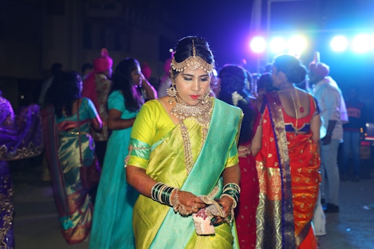 A woman in ornate traditional attire stands with a contemplative expression among a group of people at a night event. Her outfit includes a bright green and turquoise saree, elaborate jewelry, and henna-decorated hands. In the background, others are also dressed in colorful garments, with vibrant lights shining from above creating a festive atmosphere.