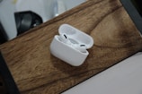 A pair of wireless earphones rests in an open charging case on a wooden surface. The earphones are white and feature a sleek, modern design. The background includes a blurred portion of another object, possibly a part of a desk or appliance.