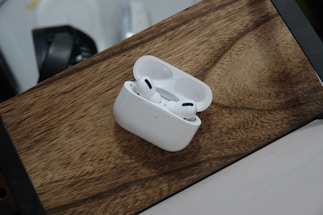A pair of wireless earphones rests in an open charging case on a wooden surface. The earphones are white and feature a sleek, modern design. The background includes a blurred portion of another object, possibly a part of a desk or appliance.