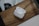 A pair of wireless earphones rests in an open charging case on a wooden surface. The earphones are white and feature a sleek, modern design. The background includes a blurred portion of another object, possibly a part of a desk or appliance.