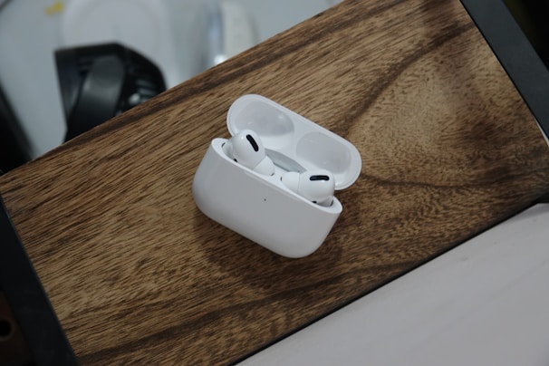 A pair of wireless earphones rests in an open charging case on a wooden surface. The earphones are white and feature a sleek, modern design. The background includes a blurred portion of another object, possibly a part of a desk or appliance.