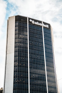 A tall, modern skyscraper with a grid of reflective windows and a prominent logo at the top. The building has a sleek design with vertical lines accentuating its height. The sky is partly cloudy, adding to the urban atmosphere.