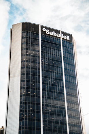 A tall, modern skyscraper with a grid of reflective windows and a prominent logo at the top. The building has a sleek design with vertical lines accentuating its height. The sky is partly cloudy, adding to the urban atmosphere.